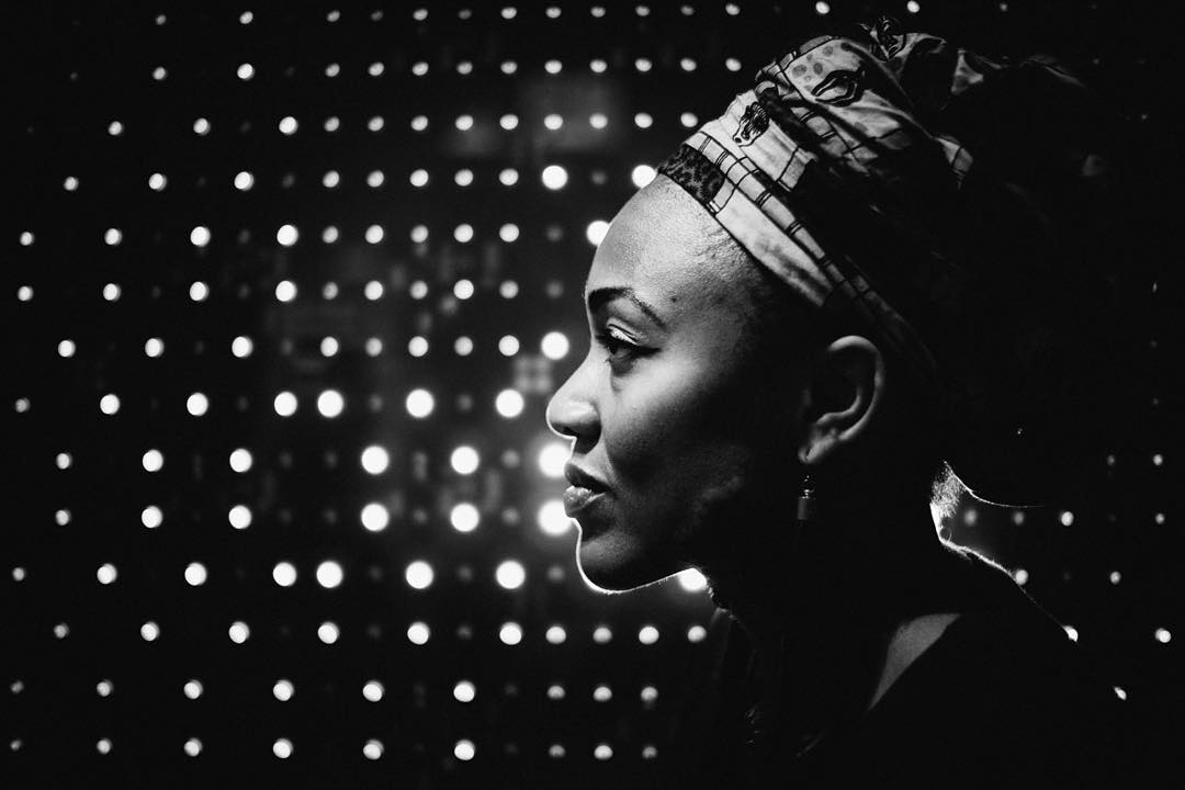 Profile portrait of a woman in headwrap, softly lit against a field of glowing dots in shadowed black and white.